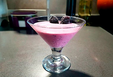 Imagen de un postre cremoso púrpura en copa de cóctel, decorado con dos moras. Fondo de cocina.