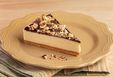 Imagen de una porción de cheesecake con cobertura de chocolate y trozos crujientes en un plato amarillo.