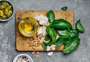Imagen de los ingredientes para el pesto: aceite de oliva, albahaca, nueces, ajo, queso y aceitunas, sobre una tabla de madera.