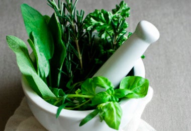 Imagen de un mortero y una mano de mortero blancos llenos de hierbas frescas como romero, salvia, albahaca y perejil.