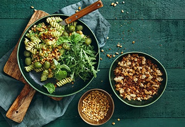 Imagen de un plato de pasta fusilli verde con pesto, aceitunas, rúcula, piñones y pan rallado crujiente sobre una mesa verde oscuro.
