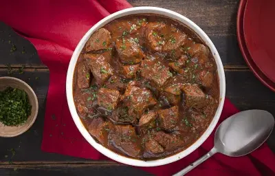 Fotografia em tons de marrom e vermelho, tendo ao centro uma panela com ensopado de carne e molho de cerveja preta, sobre guardanapo vermelho, ao lado um potinho com salsinha picada, uma colher de servir e uma tampa. Tudo sobre bancada de madeira rústica. Fotografia em tons de marrom e vermelho, tendo ao centro uma panela com ensopado de carne e molho de cerveja preta, sobre guardanapo vermelho, ao lado um potinho com salsinha picada, uma colher de servir e uma tampa. Tudo sobre bancada de madeira rústica.