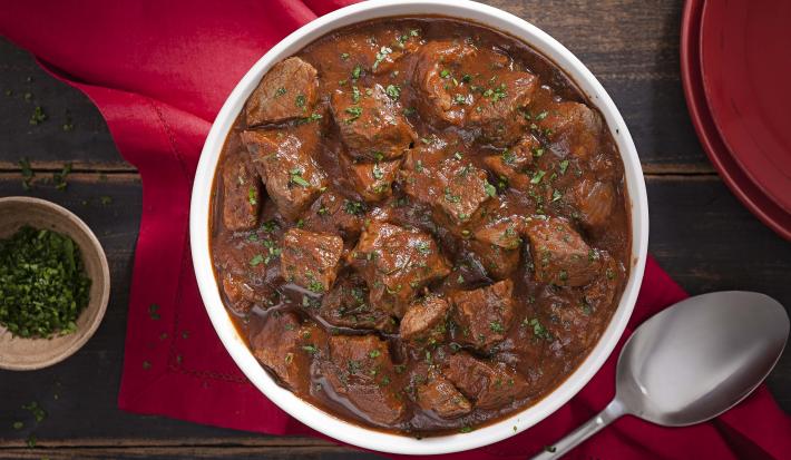 Fotografia em tons de marrom e vermelho, tendo ao centro uma panela com ensopado de carne e molho de cerveja preta, sobre guardanapo vermelho, ao lado um potinho com salsinha picada, uma colher de servir e uma tampa. Tudo sobre bancada de madeira rústica.