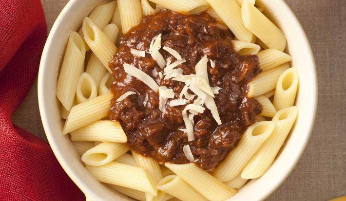 Fotografia em tons de amarelo e vermelho, ao centro uma panela destampada com o macarrão tipo penne e o ragú em cima decorado com queijo ralado. Ao lado está a tampa amarela da panela e um pote vermelho com queijo ralado.